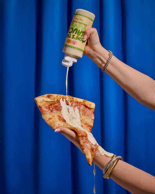 Person holding a slice of pizza, pouring a bottle label "She's the Sauce Creamy Ranch" against a blue curtain.