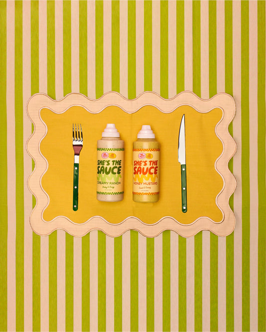 Two bottles of 'She's the Sauce' on a yellow place setting with a decorative border, against a striped green and beige background.