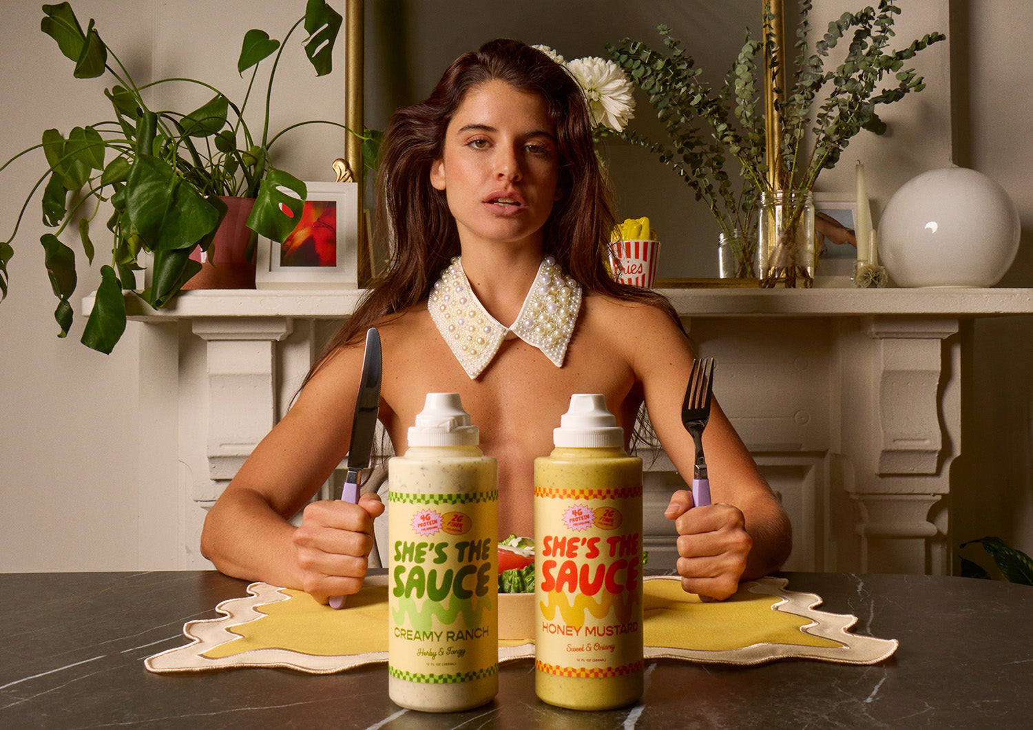 Woman sitting at a table with fork and knife in hand and two 'She's the Sauce' bottles infront of her in a home setting