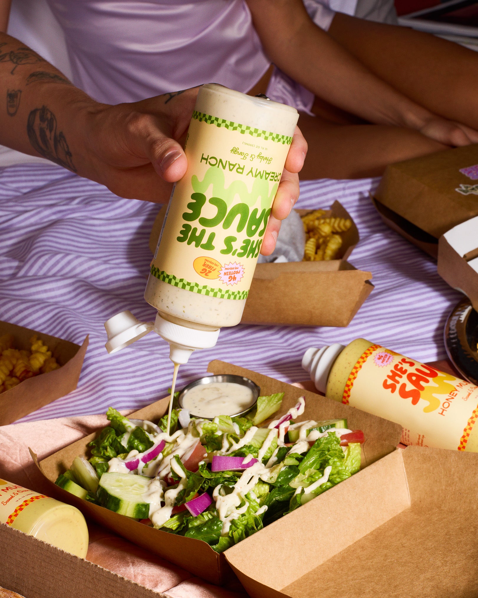 Person pouring a bottle of sauce labeled "She's the Sauce Creamy Ranch"onto a salad surrounded by other takeout containers on a purple striped bed.