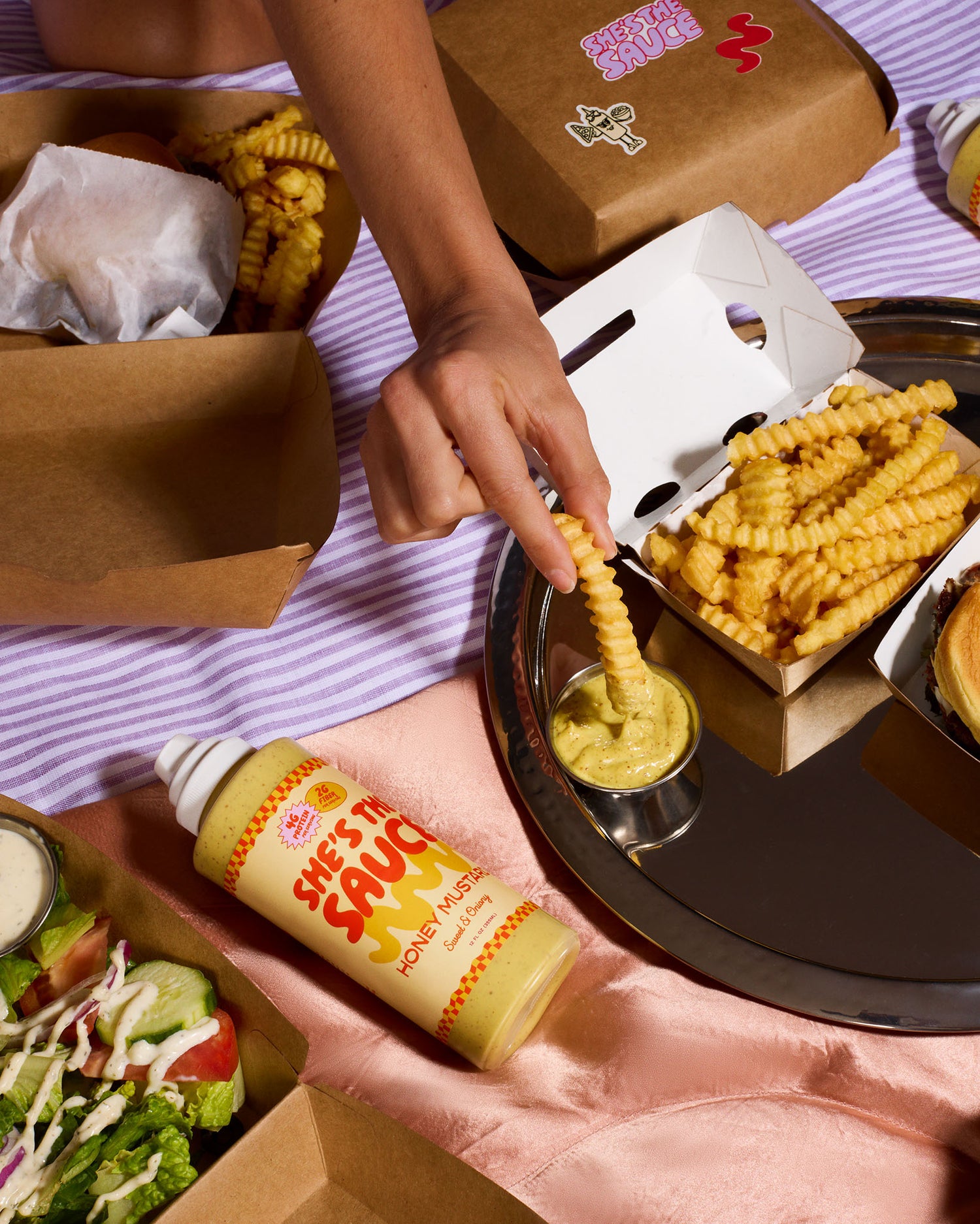 Person dipping a crinkle-cut fry into a container of She's the Sauce Honey Mustard on a mirror tray.