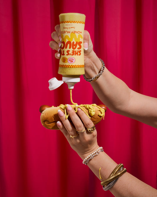 Person holding a hotdog, pouring a bottle label "She's the Sauce Honey Mustard" against a red curtain.
