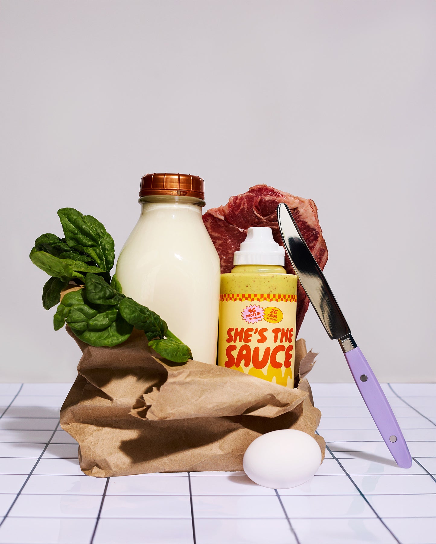 Grocery items including milk, spinach, meat, a bottle labeled 'She's the Sauce Honey Mustard', an egg, and a knife on a white surface.